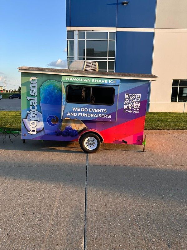 Tropical Sno Hawaiian Shave Ice trailer parked on pavement in front of a blue and white building under a clear blue sky.