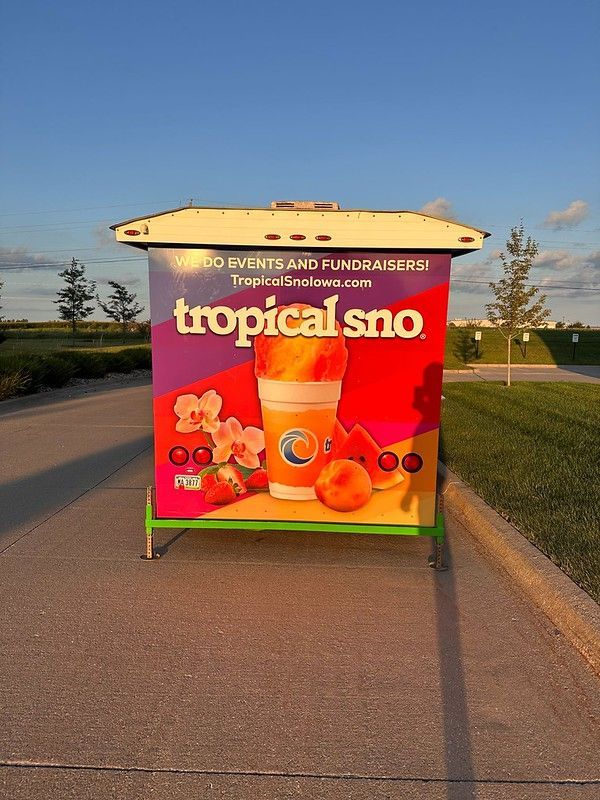 A Tropical Sno mobile kiosk with a colorful advertisement featuring a shave ice cup, fruit, and flowers at sunset.
