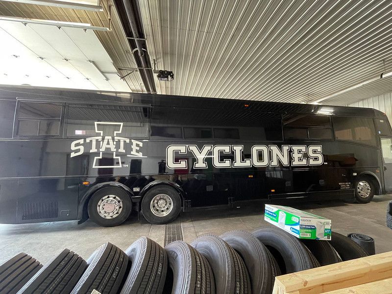 A black Iowa State Cyclones team bus parked inside a garage with several large tires in the foreground.