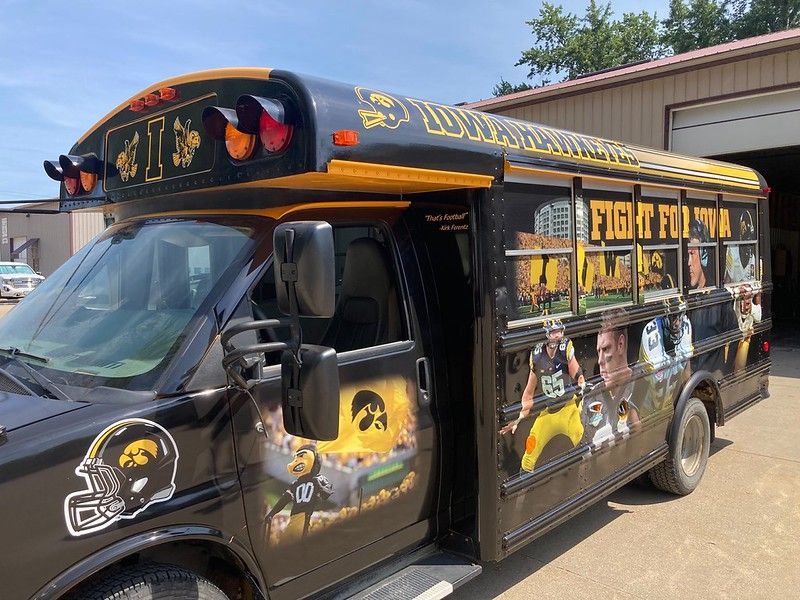 A black and gold Iowa Hawkeyes-themed bus parked outside, decorated with team logos, mascots, and sports imagery.