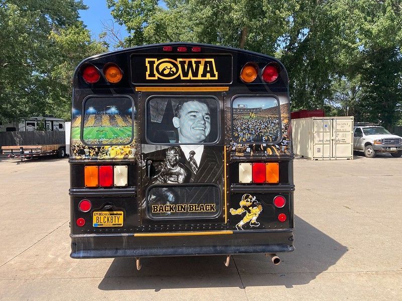 A black bus decorated with University of Iowa Hawkeyes branding, featuring stadium imagery and a portrait of a coach.