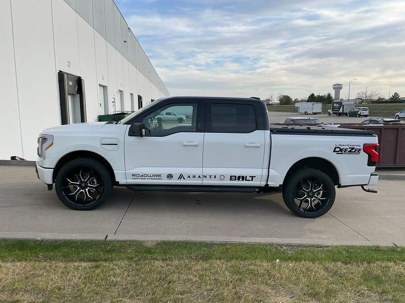 A white Ford F-150 Lightning pickup truck with custom black rims parked outside a large white warehouse building.