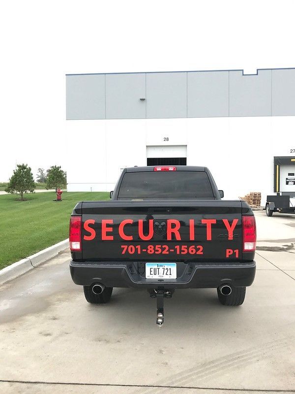 A black security pickup truck parked outside a white industrial building, displaying red lettering and a phone number.