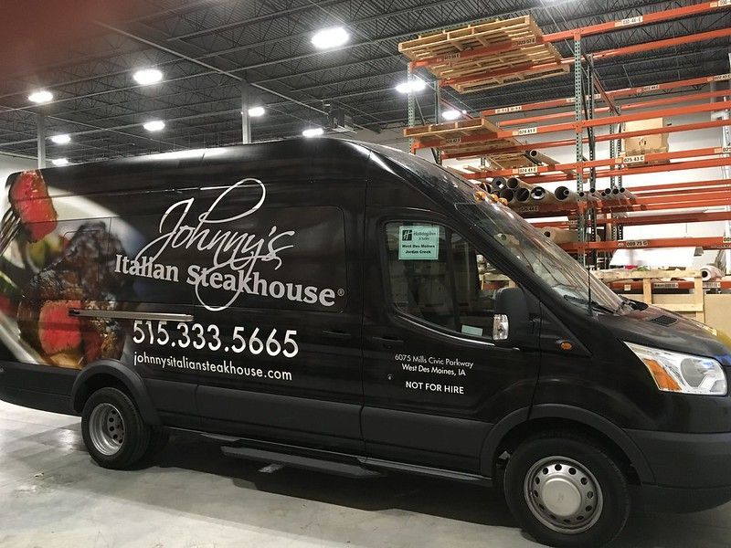A black Johnny's Italian Steakhouse delivery van parked inside a warehouse with tall storage racks.