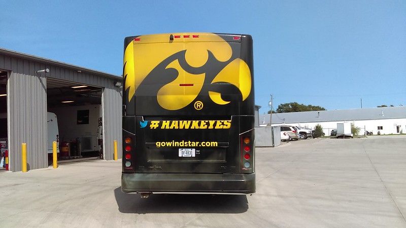 Back view of a black bus branded with a large yellow Iowa Hawkeyes logo, parked outside a warehouse on a sunny day.