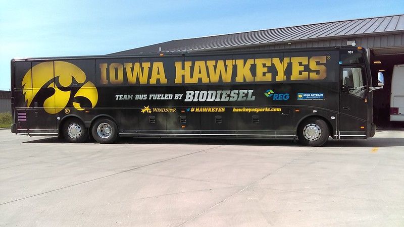 A black Iowa Hawkeyes team bus parked on a concrete lot, featuring a large yellow team logo and biodiesel branding.