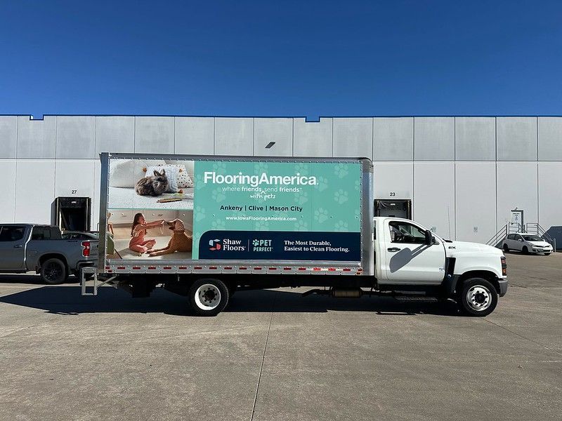 A white Flooring America delivery truck parked outside a large warehouse building.