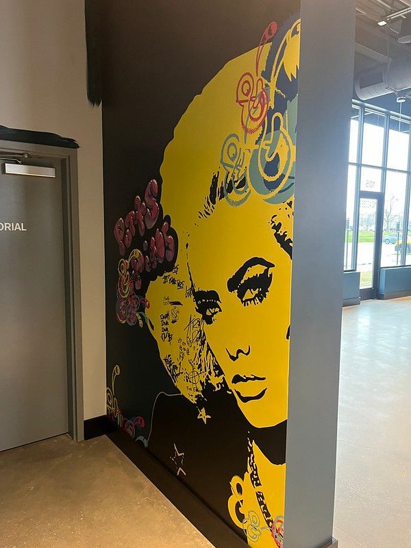 A large, bright yellow pop-art style mural of a face on a black wall next to a gray door in an open building.