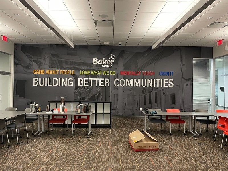 A modern office meeting room with tables and chairs in front of a wall mural featuring the Baker Group logo and mission.