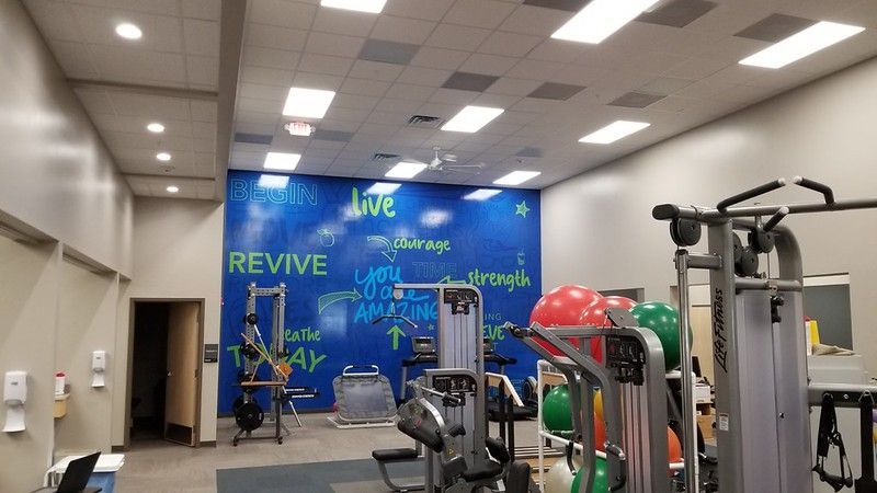 Gym interior with weight machines, exercise balls, and a blue wall featuring inspirational text.