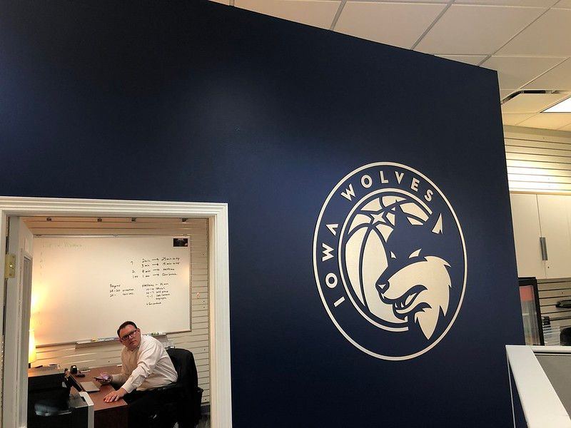 A person sits at a desk inside a small office with a large Iowa Wolves basketball logo on the adjacent navy wall.