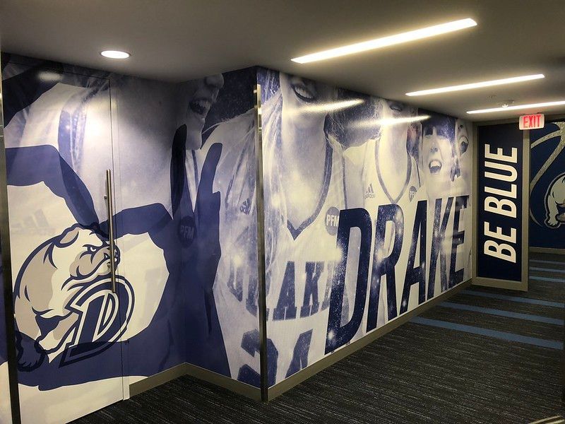 Blue and white wall mural featuring Drake University branding, bulldog mascot, and players, lining an indoor hallway.