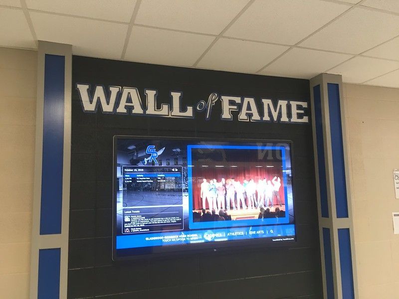 A Wall of Fame sign hangs on a dark brick wall above a television screen displaying a group photograph.