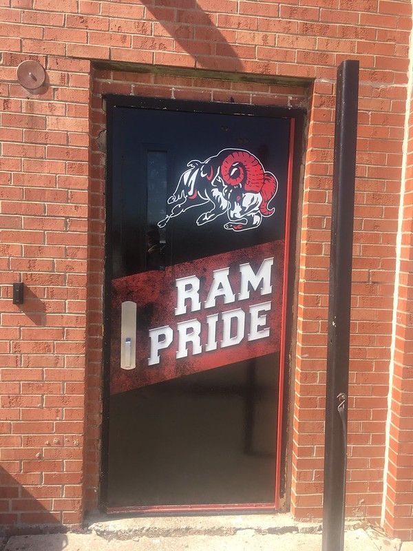 A brick-walled entrance features a black glass door with a white ram mascot and the text 