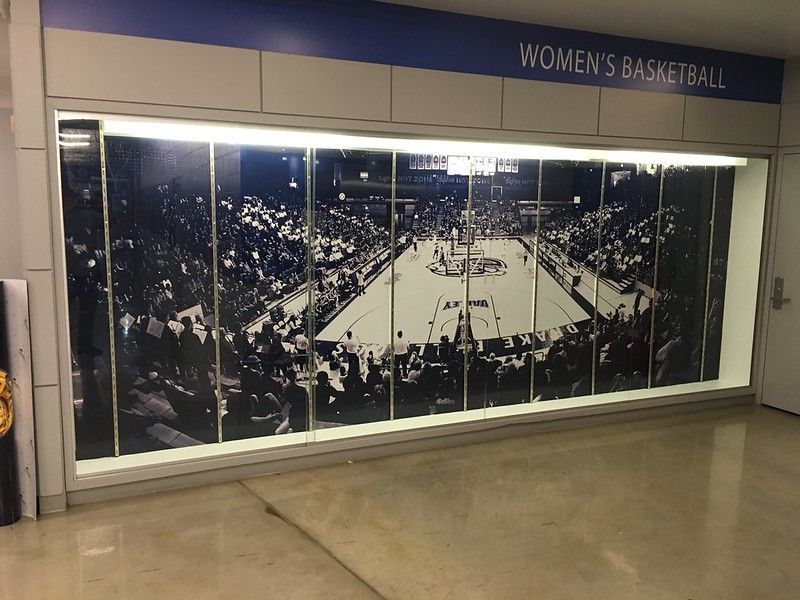 A wide, backlit wall display showing an overhead view of a basketball court during a game under the Women's Basketball sign.