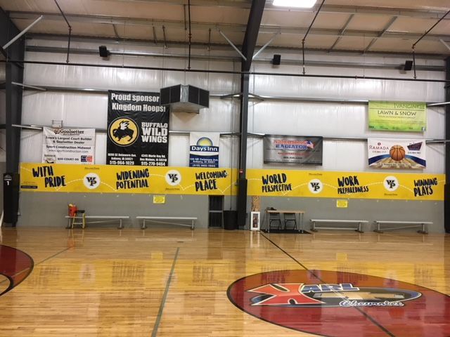 Indoor basketball court with a yellow banner featuring text, sponsor signs on the wall, and a logo on the hardwood floor.