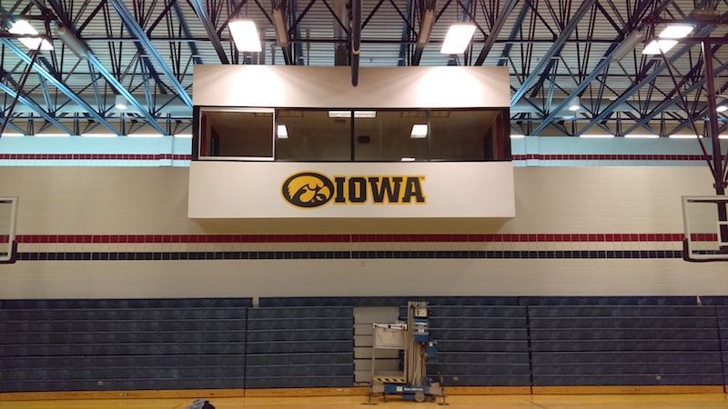 A high-angle view of a gymnasium featuring a wall-mounted Iowa Hawkeyes logo sign below a windowed viewing room.