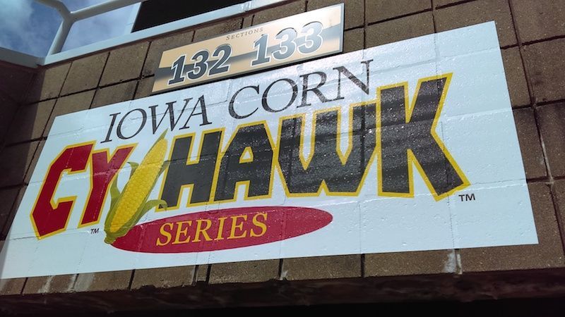 Sign for the Iowa Corn Cy-Hawk Series, featuring a corn icon, mounted on a stadium wall above seating sections 132-133.