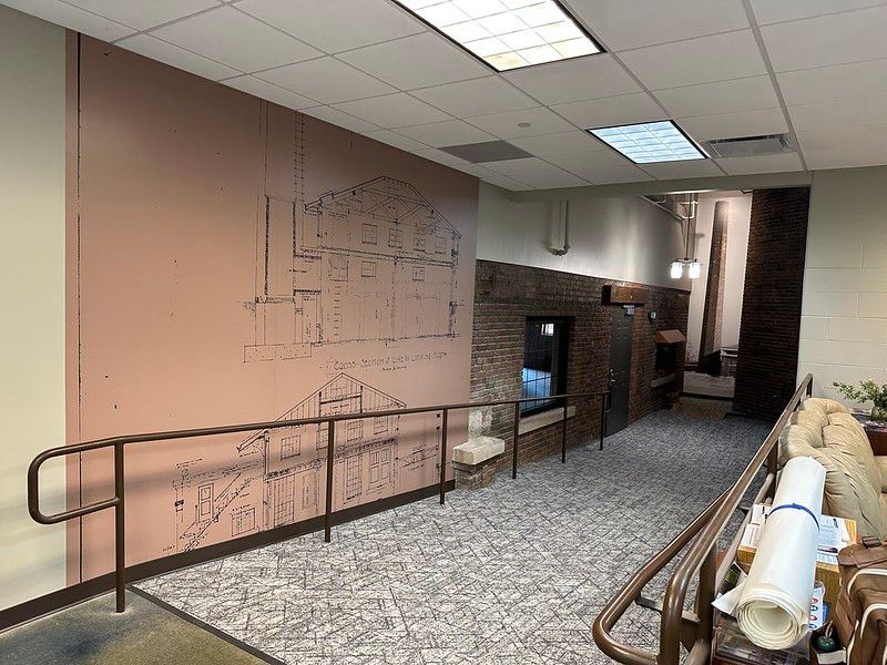 A hallway with a beige wall featuring architectural line drawings, a metal handrail, brick accents, and tiled flooring.