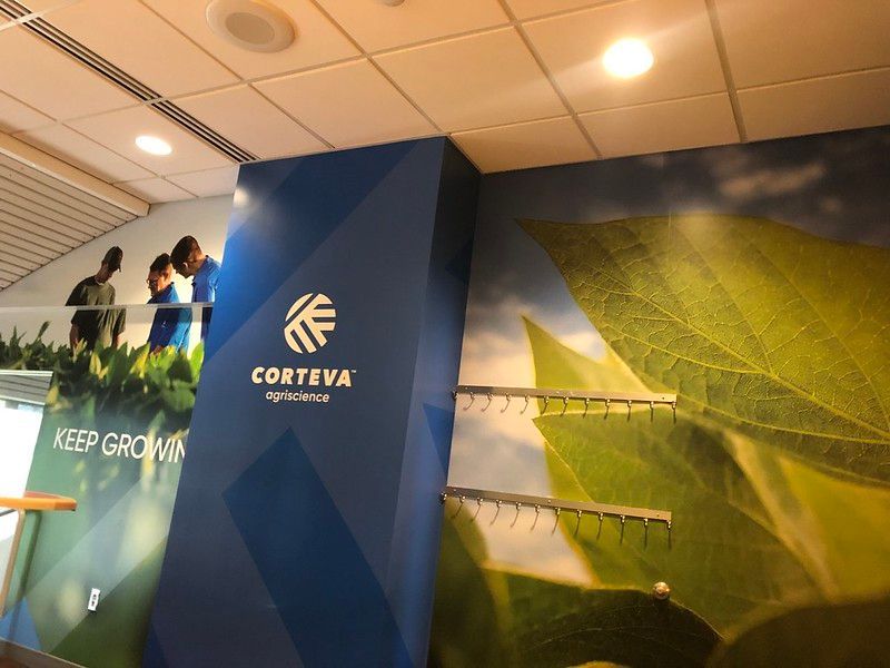 A blue wall featuring the Corteva Agriscience logo, next to a wall mural of green leaves and coat hooks.