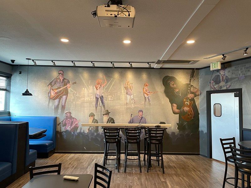 Interior of a restaurant featuring a large wall mural of rock musicians, bar seating, and tables on a wood floor.