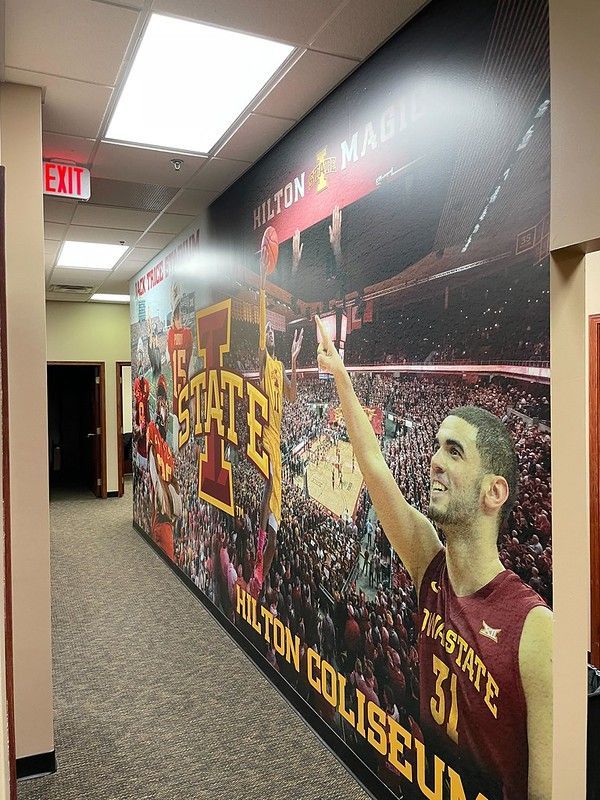 A hallway wall mural features a basketball player in a cardinal jersey celebrating in a crowded Hilton Coliseum.