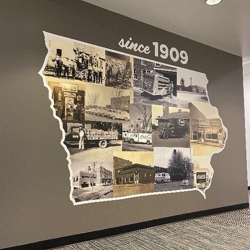 A wall mural shaped like Iowa filled with black-and-white photos of historical Coca-Cola vehicles and buildings.