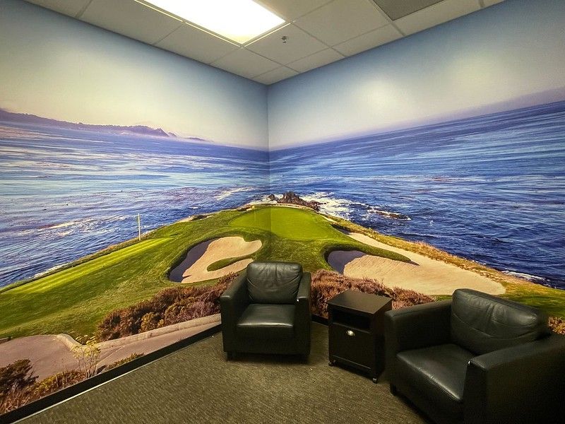A room with two black leather chairs, a small side table, and a wall mural of a coastal golf course.