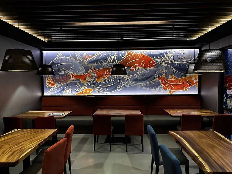 A modern restaurant interior featuring wooden tables, dark chairs, a long banquette, and a backlit mural of swimming fish.