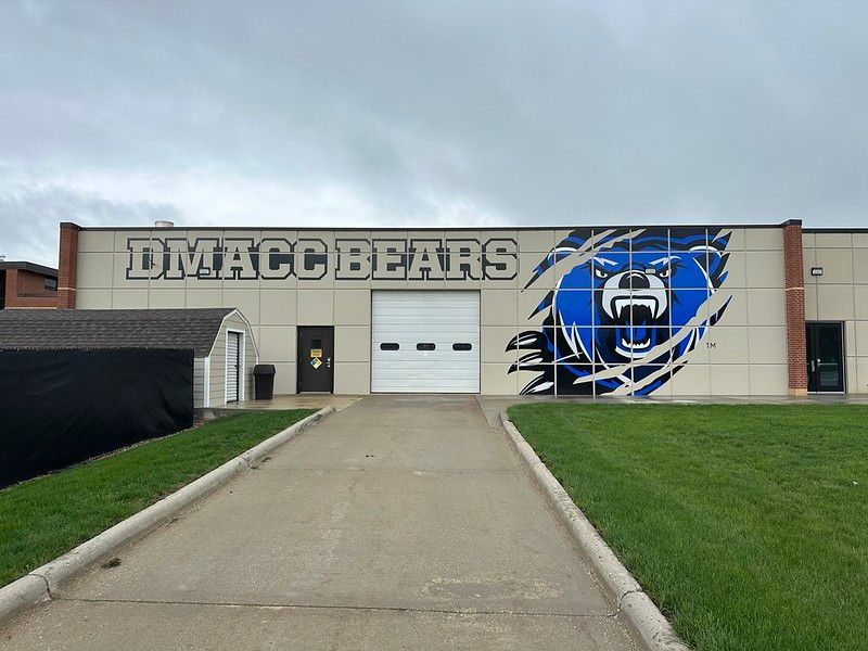 DMACC Bears sign and a large blue bear graphic on the wall of a building with a center garage door and grassy lawn.