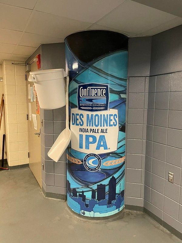 A pillar wrapped in a Confluence Brewing Co. Des Moines IPA sign, with a wall-mounted bucket and scoop beside it.