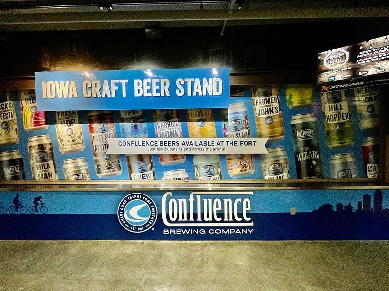 A blue Iowa Craft Beer Stand kiosk with Confluence Brewing Company branding and a background of various beer cans.