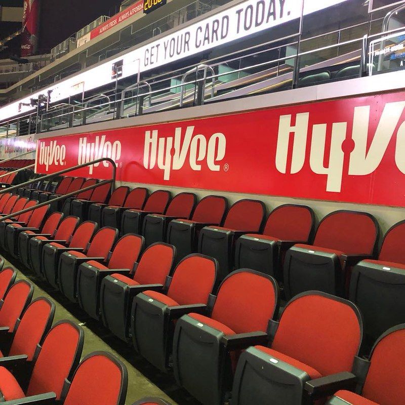Empty red stadium seats in rows with Hy-Vee advertisements on the wall behind them.