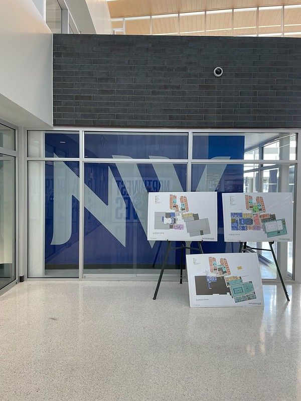 Three floor plan posters on easels stand in a bright, modern building lobby with a large blue 