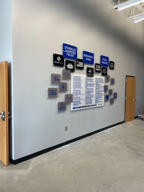 Donor wall with blue and black plaques surrounding a list of names on a light gray wall in an office hallway.