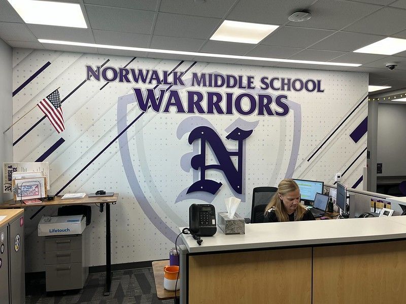 An administrative office reception area with a Norwalk Middle School Warriors wall graphic and a staff member working.
