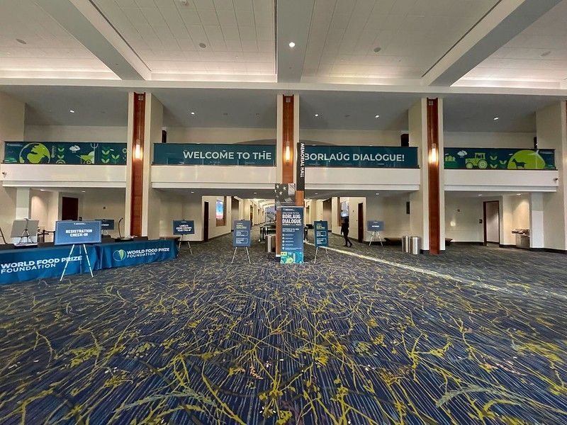 A wide, empty convention center hallway with patterned blue carpet, banners for the Borlaug Dialogue, and registration desks.