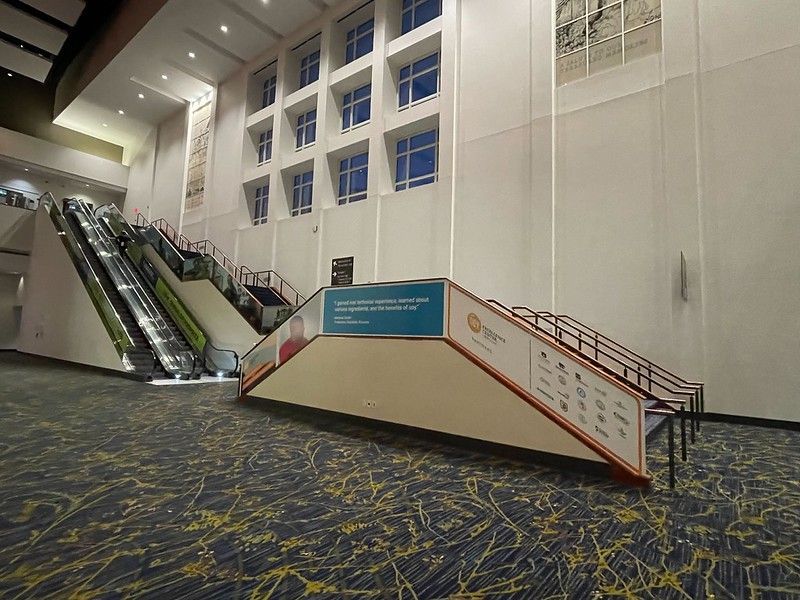 A modern lobby with an escalator, a staircase with event branding on its side, and patterned carpet under high ceilings.