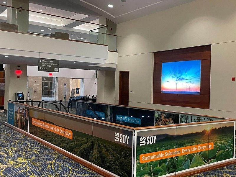 An indoor lobby features a balcony railing wrapped in US Soy promotional signage depicting lush green soybean fields.