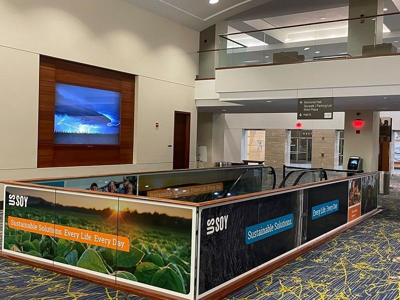 A brightly lit indoor building lobby featuring a prominent escalator enclosed by branded promotional wall panels.