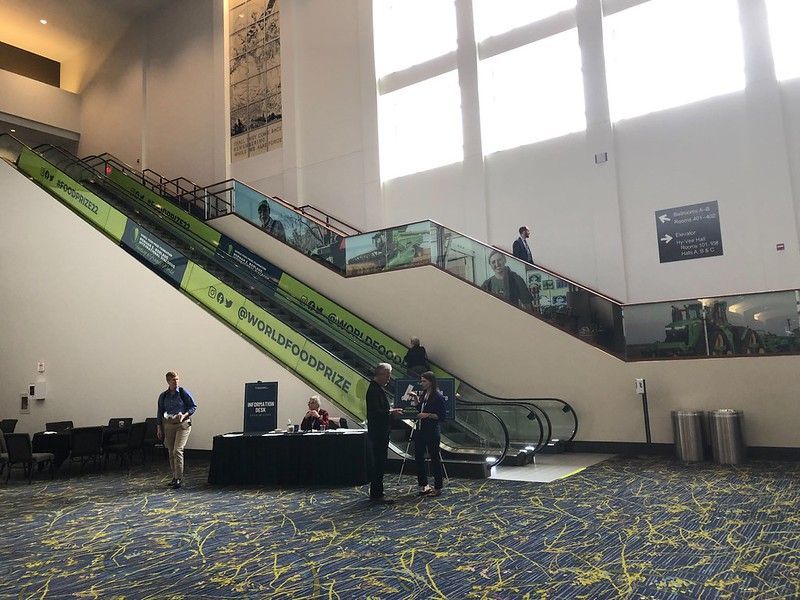 People stand near branding-covered escalators in a high-ceilinged hotel or convention center lobby.