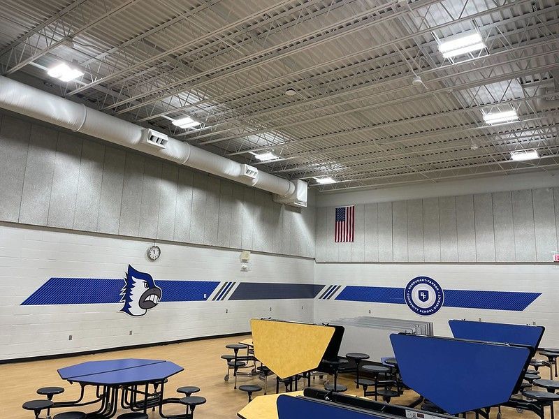 A school gymnasium or cafeteria with blue and white wall graphics, folding tables, and industrial ceiling lights.