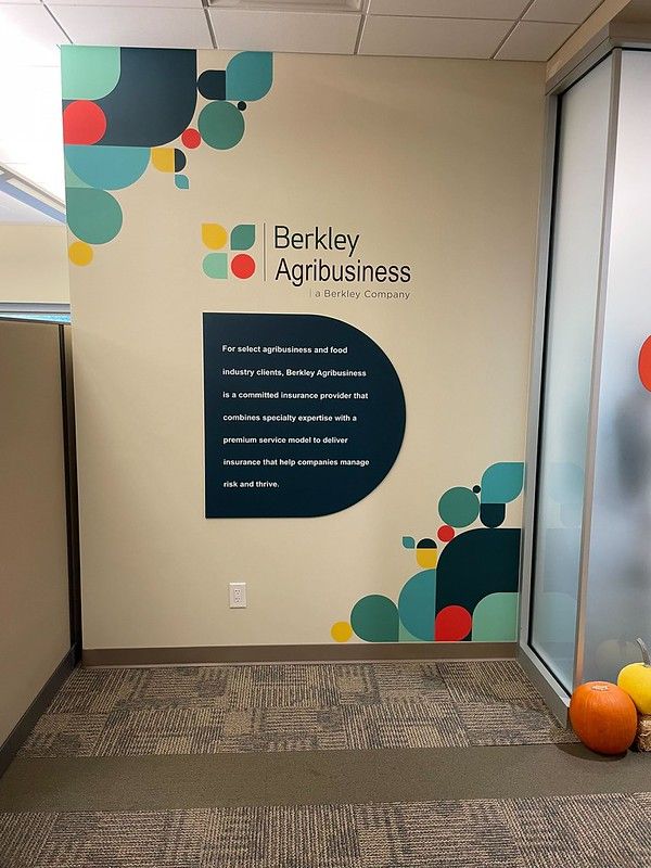 An office wall with a Berkley Agribusiness sign and logo, featuring blue and orange geometric shapes and two pumpkins.
