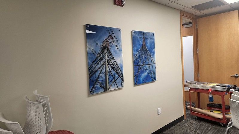 Two rectangular artworks depicting electrical transmission towers hang side-by-side on a beige wall in an office setting.
