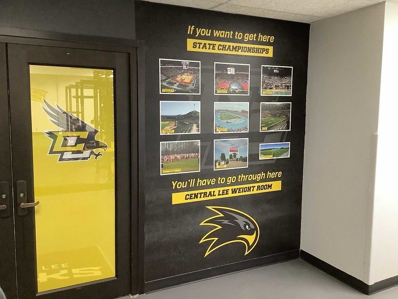 A gym entrance with a door featuring a hawk logo, and a black wall with a collage of stadium photos and motivational text.