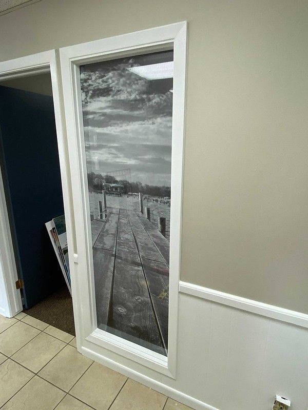 A window with a black-and-white print of a wooden dock extending over water on a wall with beige and white trim.