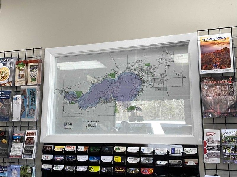 A framed map of Clear Lake, Iowa, mounted on a wall above a display rack of brochures in a travel information center.