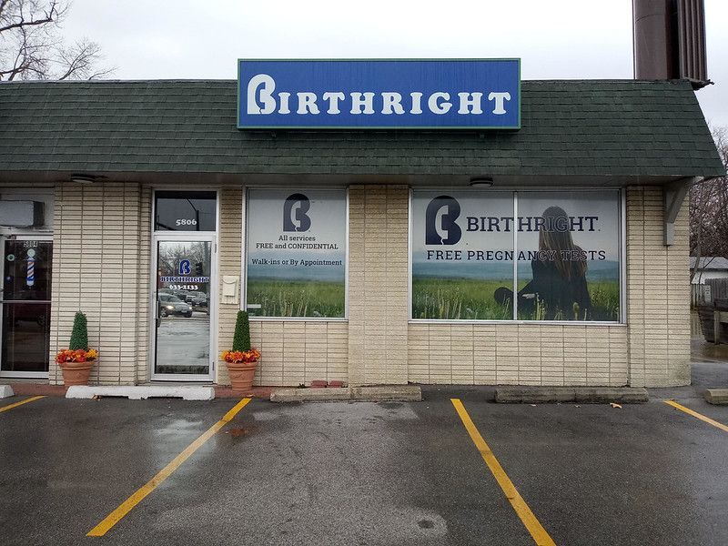 The storefront of a Birthright center with a blue sign, white brick facade, glass doors, and parking spaces in front.