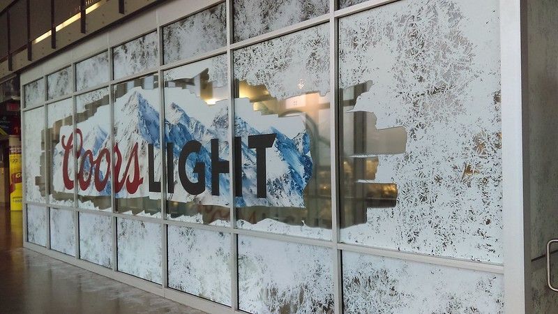 A Coors Light advertisement on a glass store window with a graphic of blue mountain peaks and frosted glass patterns.