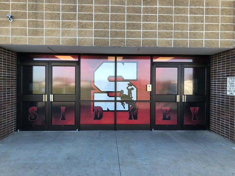 Exterior doors of Sidney school building with a large red “S” logo and the word “SIDNEY” displayed on glass panes.